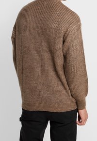 Man som bär en brun grovstickad tröja och svarta byxor, sedd bakifrån mot en enkel ljusbakgrund.