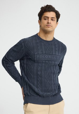 Uomo con capelli scuri e ricci che indossa un maglione blu navy a trecce con la scritta "Polo Club Established 2019", in piedi con le mani dietro la schiena.