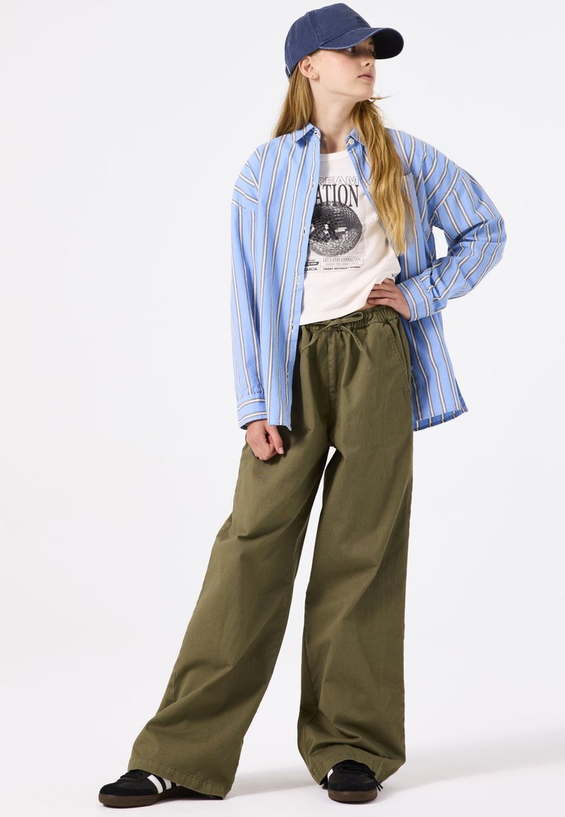 Ein blau-weiß gestreiftes Button-Up-Hemd über einem Grafik-T-Shirt, kombiniert mit lockeren olivgrünen Hosen und schwarzen Sneakers, abgerundet mit einer Jeansmütze.