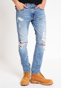 Hombre vistiendo jeans rasgados de color azul claro con las manos en los bolsillos y botas de trabajo de color beige con cordones, sobre fondo blanco.