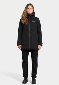 Jovem mulher em pé, ereta, vestindo uma jaqueta preta com capuz e fecho éclair, calças pretas e botas pretas com atacadores, num fundo branco.