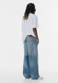 Prevelik sivi bombažni majica v kombinaciji z svetlo modrimi širokimi jeans hlačami, ki imajo rjavo usnjeno oznako na hrbtni strani.