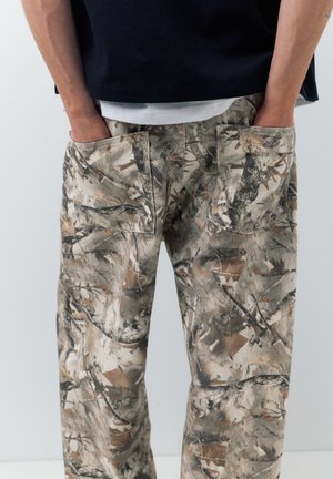 Ruky v zadných vreckách voľných nohavíc s maskáčovým vzorom, nosené s čiernou košeľou na bielom tričku.