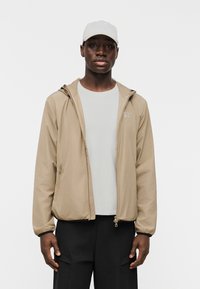 Άνδρας που φοράει μπεζ φούτερ με κουκούλα και φερμουάρ πάνω από λευκό πουκάμισο, μαύρο παντελόνι και λευκό καπέλο μπέιζμπολ, στέκεται μπροστά σε λευκό φόντο.