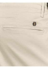 Beige stof close-up met een achterzak die een slanke horizontale gleuf heeft en een kleine zwarte hexagonale knop erboven.