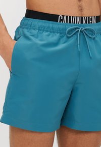 Τελειόμορφες γαλαζοπράσινες μαγιώ με ελαστική μέση, μαύρο λογότυπο Calvin Klein, πλαϊνές τσέπες και κορδόνι για ρυθμιζόμενη εφαρμογή. Λαμπερή υφή.