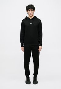 Conjunto de sudadera con capucha y pantalones negros en tela suave; la sudadera con capucha incluye cordones y logo EA7; diseño ajustado con puños y tobillos acanalados; zapatillas negras.