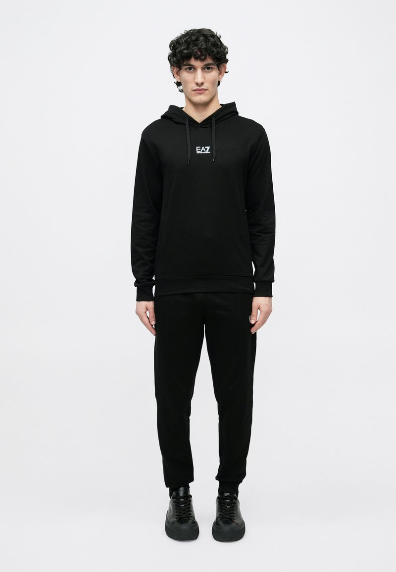 Conjunto de sudadera con capucha y pantalones negros en tela suave; la sudadera con capucha incluye cordones y logo EA7; diseño ajustado con puños y tobillos acanalados; zapatillas negras.
