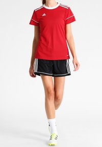 Röd idrottströja med vita detaljer och Adidas-logga, tillsammans med svarta shorts med vita ränder. Klara gula skor och vita strumpor.