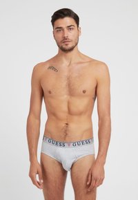 Graue Baumwollslips mit einem marineblauen und roten "GUESS"-Logo-Bund, featuring ein körperbetontes Design und glatte Textur. Modell zeigt die Vorderansicht.
