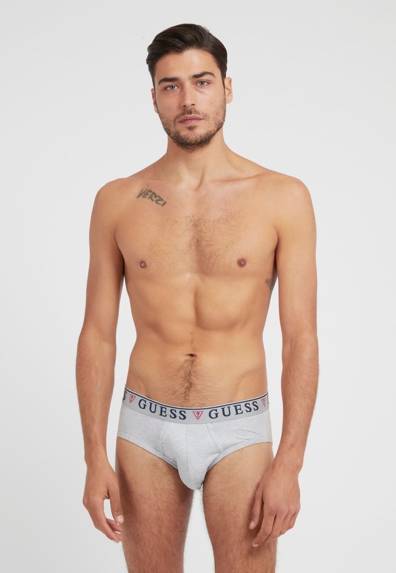 Graue Baumwollslips mit einem marineblauen und roten "GUESS"-Logo-Bund, featuring ein körperbetontes Design und glatte Textur. Modell zeigt die Vorderansicht.