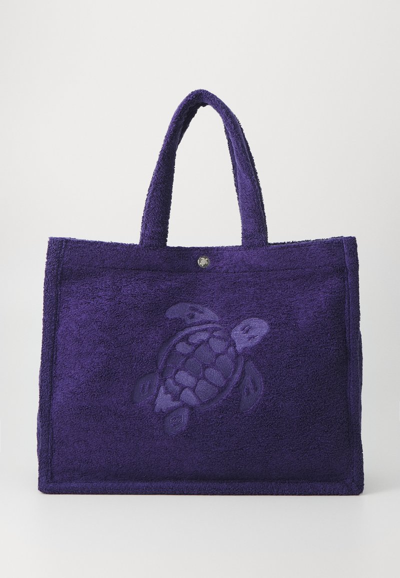 Lila Handtasche im Handtuch-Stil aus strukturiertem Stoff, mit einem geprägten Schildkrötenmotiv in der Mitte und zwei stabilen Griffen.