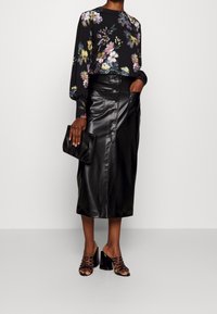 Schwarzes Blusentop mit langen Ärmeln und bunten Blumenmuster, kombiniert mit einem schwarzen Leder-Midikleid mit Frontknöpfen und Taschen. Schwarze Blockabsatzschuhe.