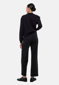 Sweatshirt en maille noire avec des poignets côtelés, associé à un pantalon large taille haute noir. Modèle portant des chaussures en cuir noir.