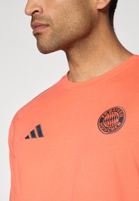Koral kortermet t-skjorte med rund hals, FC Bayern München-logo og svarte Adidas-striper på venstre skulder. Teksturert bomullsstoff.