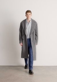 Γκρι μάλλινο παλτό με διπλό πέτο, φορεμένο πάνω από πουκάμισο με λευκές και μπλε ρίγες και μπλε παντελόνι, συμπληρωμένο με μαύρα παπούτσια.