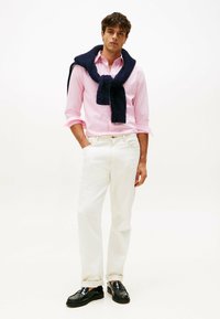 Herenoutfit met een lichtroze gestreept shirt, witte broek en een marineblauwe trui nonchalant om de nek gedrapeerd. Zwarte instappers ronden de look af.