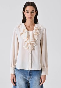 Donna che indossa una blusa avorio con rouches a strati al colletto e chiusura a bottoni, abbinata a jeans in denim blu.