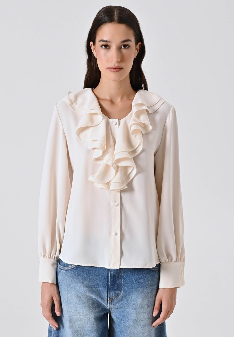 Donna che indossa una blusa avorio con rouches a strati al colletto e chiusura a bottoni, abbinata a jeans in denim blu.