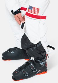 Pantalones de nieve blancos con detalles en rojo y una bandera estadounidense, puños ajustables, combinados con botas de esquí negras con suelas naranjas y múltiples hebillas.