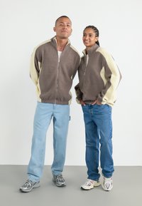 Twee modellen dragen fleece zip-up jassen, de ene in bruin met crème accenten, de andere in crème met bruine accenten, gecombineerd met spijkerbroeken en sneakers.