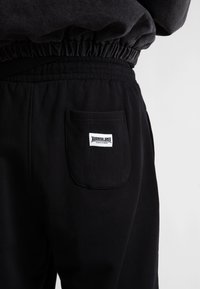 Primer plano de pantalones de chándal negros con cinturilla elástica y un bolsillo trasero que presenta una etiqueta blanca que dice "Wasted Always Faithful."
