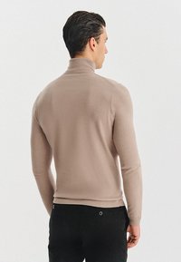 Jasnobrązowy sweter z golfem wykonany z miękkiego dzianinowego materiału, o dopasowanym kroju z prążkowanym kołnierzem i brzegiem. Noszony z czarnymi spodniami.