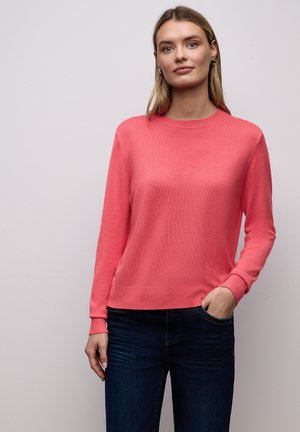 Heller Korallenpullover aus weichem Strickstoff mit rundem Ausschnitt und gerippten Bündchen. Kombiniert mit dunklen Jeans, glatte Textur sichtbar.