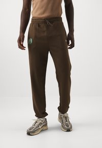 Pantaloni della tuta marroni con vita regolabile, tasche laterali e ricamo di un logo verde sulla gamba sinistra. Tessuto testurizzato e vestibilità aderente.