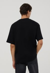 Maglietta in cotone nera con vestibilità comoda, maniche corte e scollo rotondo. La vista posteriore mostra la tessitura del tessuto liscia e un design minimalista.