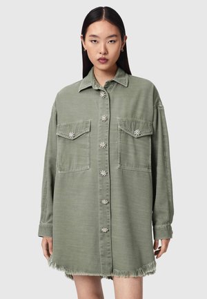 Vrouw draagt een oversized olijfgroen overhemd met twee borstzakken en een rafelige zoom, staand tegen een effen lichte achtergrond.