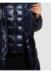 Tamsiai mėlyna pūkinė striukė blizgiu paviršiumi, su dygsniuotomis dalimis, dviem užtrauktukais ir juoda apdaila kraštuose.