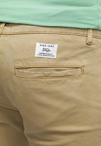 Khaki kalhoty s zadní kapsou, na které je štítek s nápisem "PIER ONE COMFORT & QUALITY PREMIUM." Hladká textura a přizpůsobený střih.
