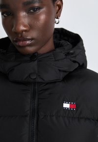 Svart dunjakke med høy krage, glidelås og to svarte trykknapper. Har et lite Tommy Jeans logo-patch på brystet.