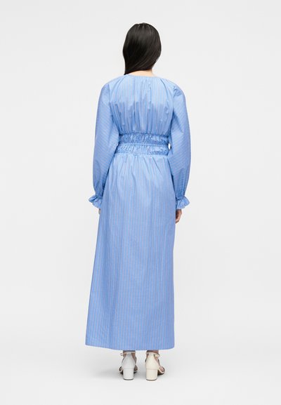 Γυναίκα με μακριά σκούρα μαλλιά που φοράει ένα μακρυμάνικο, μακρύ μπλε φόρεμα με διακριτικές ρίγες και ζωνάρι στη μέση, όρθια ξυπόλητη σε λευκό φόντο.