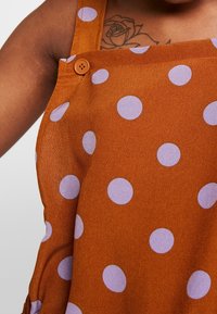 Brązowa sukienka w kropki z dużymi lawendowymi kropkami, z regulowanymi ramiączkami i guzikiem przy ramieniu. Miękka tkanina o gładkiej fakturze.