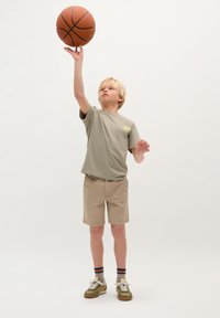 Kurzärmliges olivgrünes T-Shirt, beigefarbene Shorts und grüne Sneakers. Das Kind hält einen Basketball hoch, die gemusterten Socken sind sichtbar.