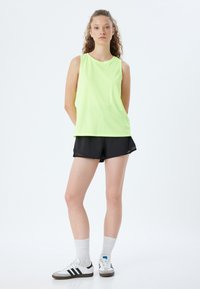 Neon zielony top bez rękawów, czarne szorty, białe sneakersy z czarnymi paskami i białe skarpetki. Gładki materiał, luźny krój, prosty design.