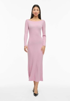 Naine seljas heleroosa, soonikkoes, pika varrukaga midi kleit ja mustad terava ninaga kontsakingad, seisab valgel taustal.