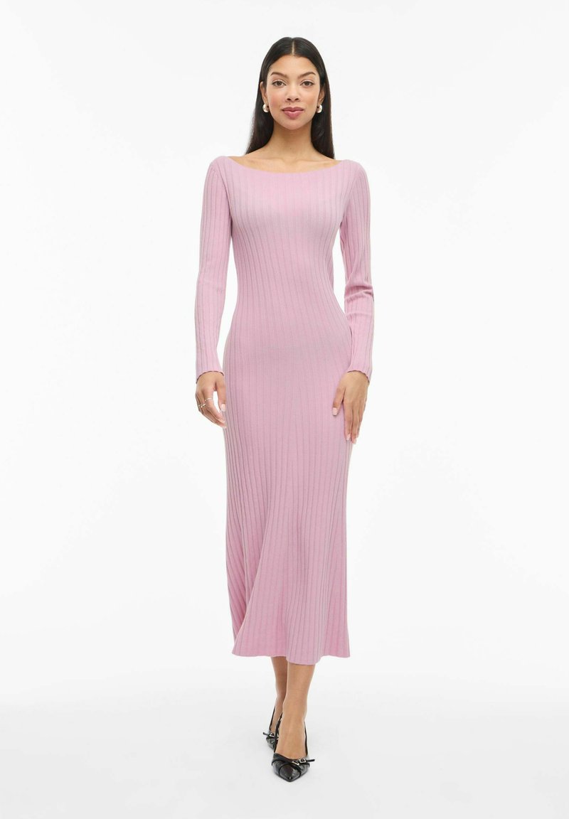 Moteris dėvi ilgomis rankovėmis, rausvos spalvos suknelę iki vidurio blauzdos su siaurais briaunomis ir juodus smailius aukštakulnius, stovinti ant balto fono.