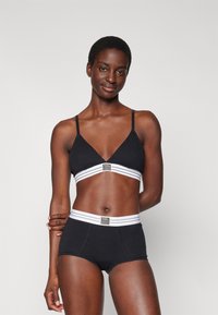 Bralette preta com alças finas e uma faixa inferior branca com riscas, combinado com calções boyshorts pretos com uma cintura de riscas brancas e etiqueta.