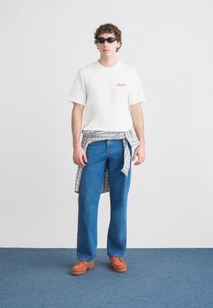 Camiseta blanca de algodón con texto rojo, vaqueros azules de mezclilla, camisa de cuadros atada a la cintura, zapatos naranjas y gafas de sol negras.