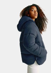 Marinblå puffer-jacka med huva, med en kviltad design och en avslappnad passform. Mjuk textur med sidofickor och knappdetaljer.