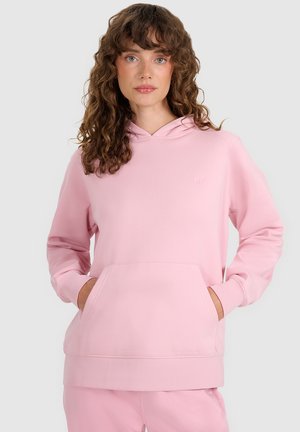 Mujer con cabello castaño rizado que lleva una sudadera con capucha rosa claro con bolsillo frontal, de pie con las manos dentro de los bolsillos contra un fondo liso.