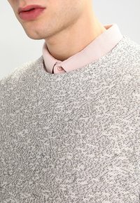 Ljusgrå strukturerad tröja med rund halsringning och en blek rosa skjorta med krage under, med två knappar och en slät yta.