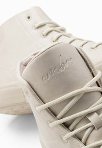 Zapatillas altas de cuero blanco con texto "ombre" grabado, cordones planos y suelas texturizadas, con un acabado suave y un diseño minimalista.