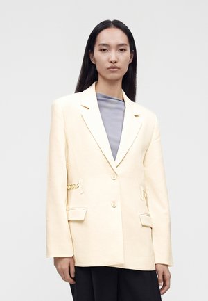 Femme aux longs cheveux noirs portant un blazer crème sur un top gris et un pantalon noir, debout devant un fond clair uni.