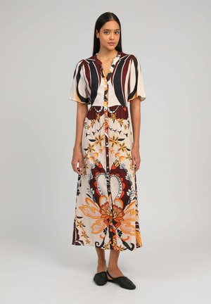 Femme debout portant une robe maxi à boutons avec des motifs abstraits orange, noir, marron et crème, et des chaussures plates noires.