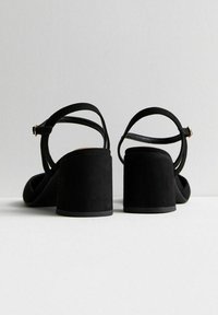 Sandali con tacco alto in suede nero, con tacchi spessi e design a fascia sottile; presentano cinturini alla caviglia regolabili e accenti hardware minimalisti.