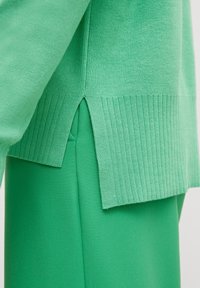 Gros plan d'un pull vert tricoté avec un ourlet côtelé et une fente latérale porté sur un pantalon vert lisse.